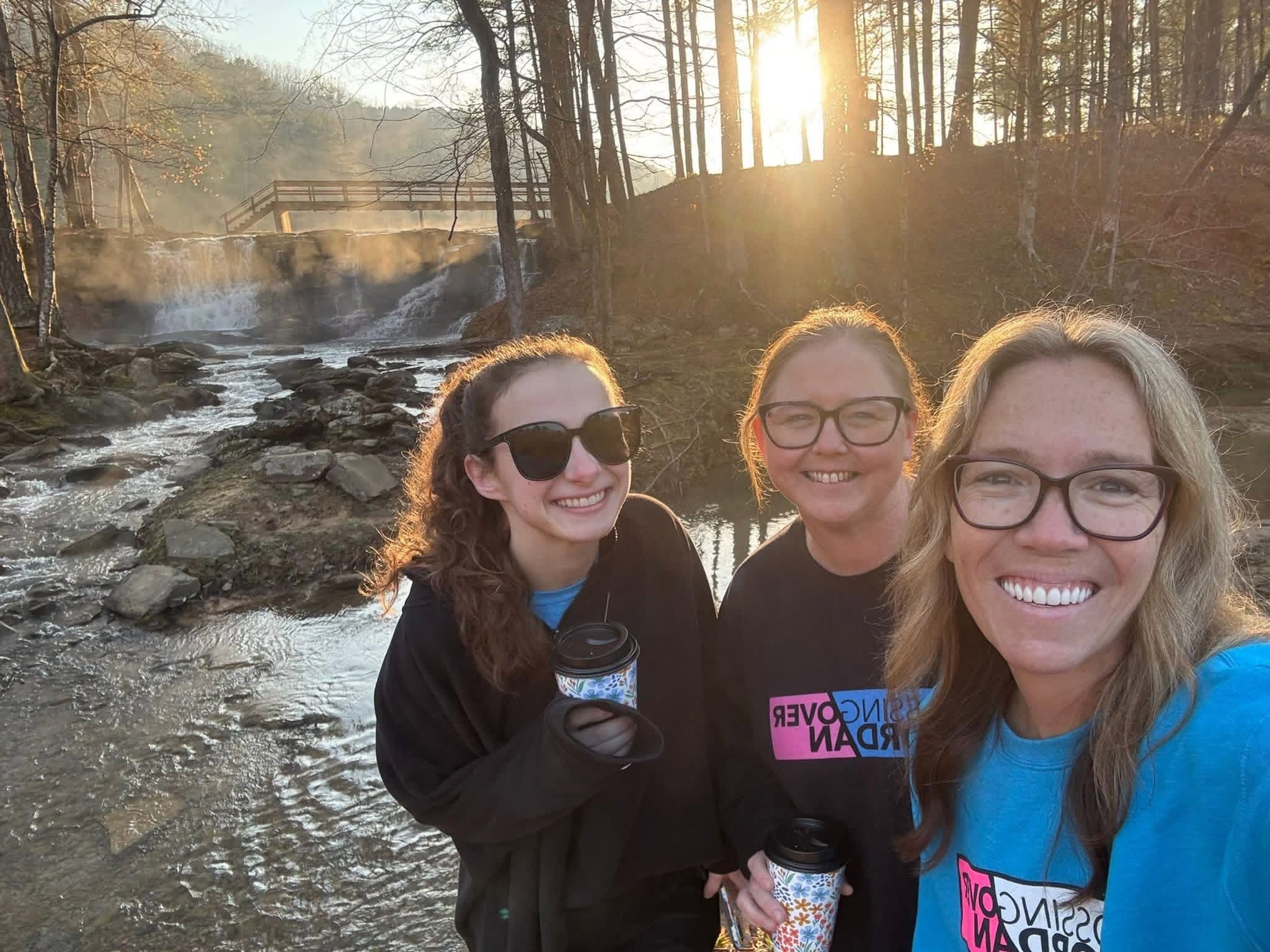 Three friends smiling together by the waterfall at sunrise during a COJ retreat